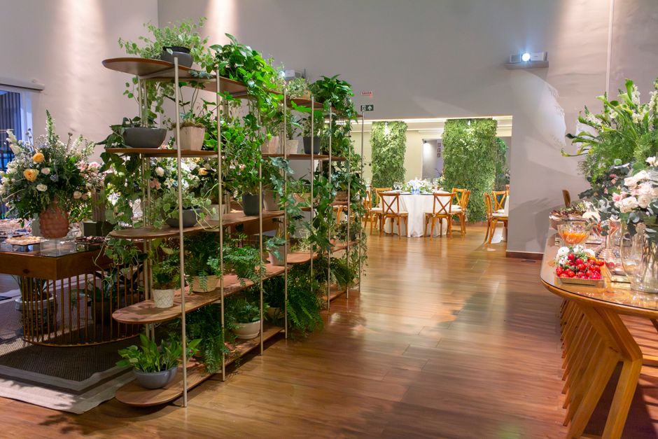 An indoor space featuring a floral display with a wooden shelving unit filled with various green potted plants and foliage, creating a lush backdrop. To the left, a large bouquet of mixed flowers, inc