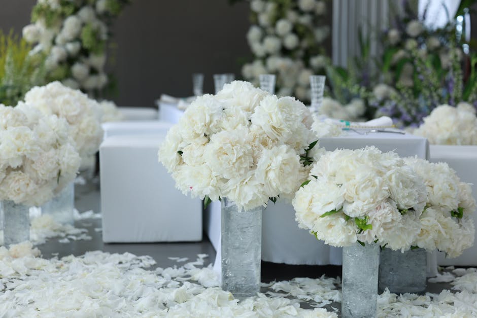 A floral arrangement featuring large, rounded bouquets of white hydrangeas and cream peonies placed in tall, rectangular glass vases. The flowers are fresh with textured, petal-rich blooms, and are se