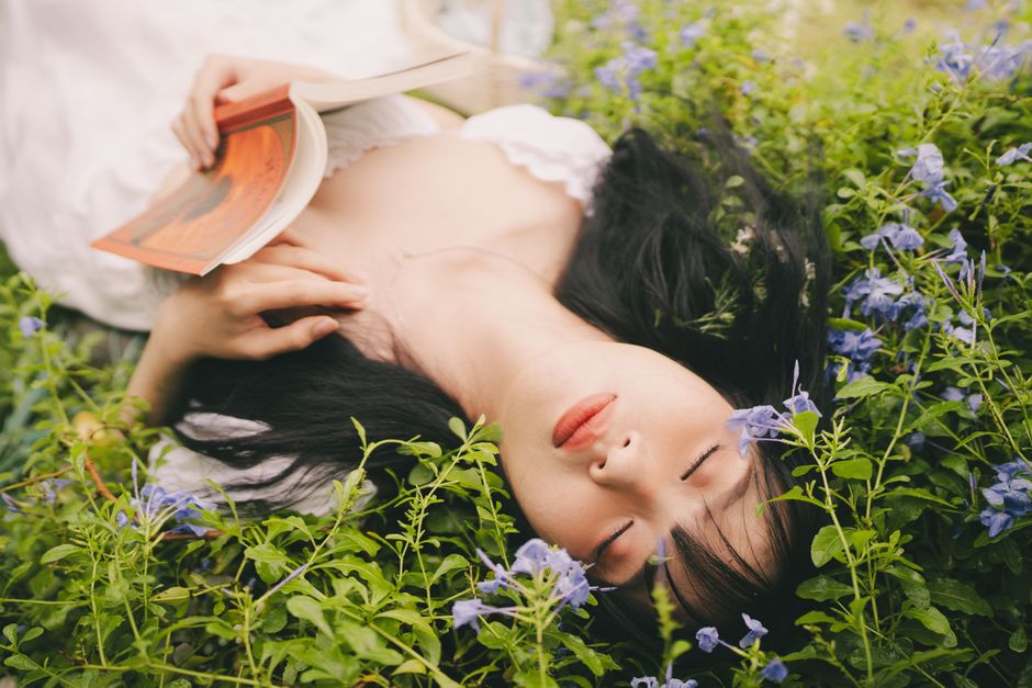 A young woman with long black hair and fair skin is lying peacefully on a bed of vibrant green foliage and purple flowers, with her eyes closed and a serene expression on her face. She is holding an o