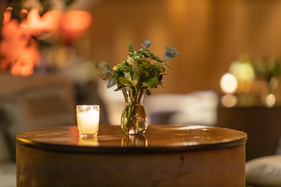 A floral arrangement in a clear glass vase featuring green hydrangeas, eucalyptus leaves, and other foliage, placed on a polished wooden table. Next to the vase is a small, textured candle holder with
