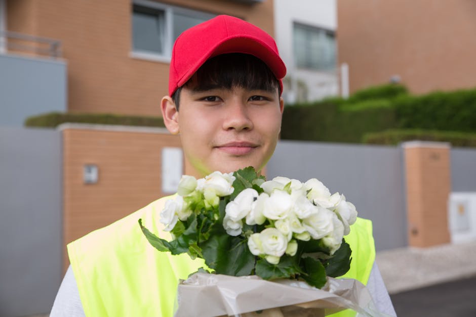 A young delivery person wearing a red cap and high-visibility yellow vest holds a bouquet of fresh white roses with green leaves, wrapped in white paper. The individual smiles subtly while standing ou