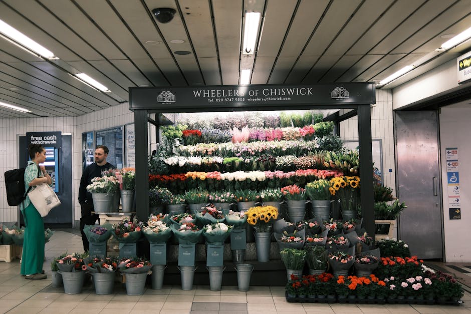 A floral display at Wheeler of Chiswick in an underground station, featuring a variety of fresh flowers arranged in multiple tiers. Brightly coloured blooms, including yellow sunflowers, pink roses, w
