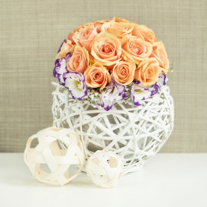 A refined floral display is carefully arranged on a smooth, reflective white surface, creating the impression of a modern wedding table set in a light-filled Vauxhall townhouse or local restaurant. At the centre sits a compact, perfectly rounded bouquet of roses in shades of soft peach, apricot, and muted salmon, each bloom open and full, with layers of velvety petals forming a gentle dome. The roses are nestled so closely together that their outlines blend, giving the top of the arrangement a plush, almost fabric-like texture. Around the lower edge of this rose sphere, a halo of white lisianthus blossoms appears, each flower edged with deep purple or violet, like delicate brushstrokes along the petal tips. These lisianthus blooms, slightly frilled and airy, break the smoothness of the dome and bring a sense of movement. Tiny tufts of white baby's breath are scattered among them, adding a misty softness that lightens the composition. The flowers emerge from a broad, rounded white basket-style container, meticulously woven from rattan or wicker into an open, lattice-like structure that shows through to the interior. This vessel has a contemporary yet handcrafted feel, echoing the artisanal pieces you might find at markets and design boutiques around Vauxhall. In front of the main arrangement rest two decorative spheres, one larger and one smaller, made from thin, light beige wooden strips woven into open frameworks. They sit casually on the tabletop, their shadows gently overlapping. Behind it all, a subtly textured beige fabric backdrop provides a warm, neutral frame, allowing the pastel blooms and natural materials to stand out. The whole scene suggests quiet celebration and timeless romance, perfect for guiding table décor choices for weddings, anniversaries, or elegant gatherings across Vauxhall.