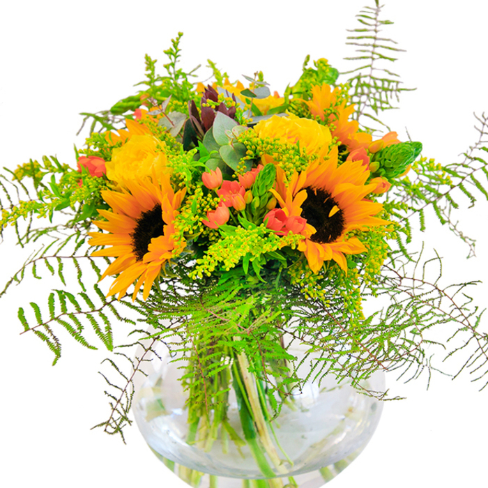 The first impression of this vibrant arrangement is one of pure, sunny exuberance, reminiscent of an afternoon spent in Vauxhall Pleasure Gardens. Towering sunflowers, their plush brown centers ringed by radiant yellow-orange petals, stand alongside plush garden roses of a soft, golden hue. Each rose gestures open in richly layered spirals, softening the display with their elegance. Polished clusters of vivid red Hypericum berries punctuate the design with unexpected cheer, while lush green ferns and light filler blooms add buoyancy and rhythm, as if caught in a passing breeze. Laced through the bouquet are slender stems of eucalyptus, their subtle blue-green leaves refreshing the composition and enhancing its wild, garden-inspired feel. The clear, spherical vase cradles the arrangement, revealing the tangle of sturdy stems as they fan out into clear water-a detail that brings a sense of grounding clarity. Gentle light dances over the bouquet, casting delicate shadows and highlighting every curve of petal and frond. The faint scent of open fields and garden blooms lingers, further enveloping the senses. Ideal for birthdays, anniversaries, or expressing heartfelt warmth, this bouquet-handcrafted by local Vauxhall florists-captures the spirit of celebration unique to this lively London neighborhood, offering a daily reminder of nature's artistry and the pleasure of giving.