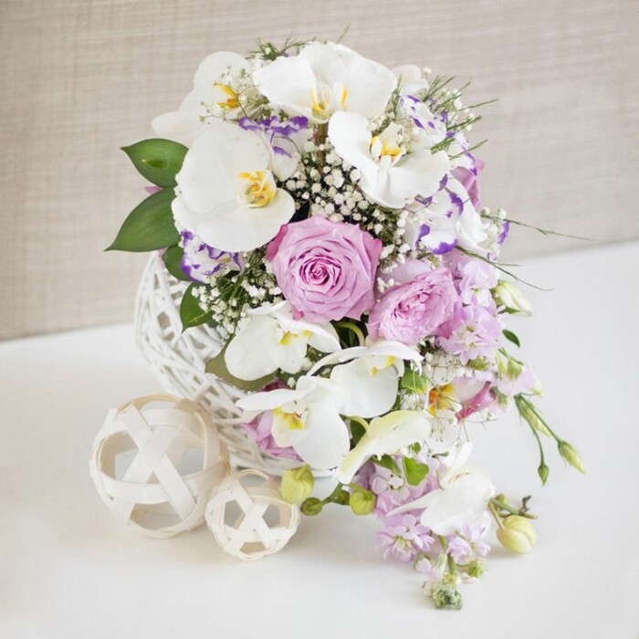 A refined wedding-style floral arrangement stands serenely on a crisp white surface, perfectly composed as if for a special celebration in one of Vauxhall's chic riverside venues. The flowers are cradled in a striking white spherical vase with an open, interlaced pattern that resembles fine wicker or sculpted rattan, its airy design allowing light to slip through and create subtle patterns around its base. Rising from this textured globe is a lush tapestry of blooms in white, blush pink, and soft purples. Several large white Phalaenopsis orchids stretch outward, their broad, curved petals gleaming like porcelain, each one centered with a delicate flare of yellow and amber that adds warmth and depth. Surrounding the orchids are plump roses in pastel pink and lavender, their layered petals spiraling inward, some fully open and others beginning to unfurl, giving the arrangement a sense of life and unfolding romance. Interspersed among them are smaller, frilled blossoms with white petals lightly brushed at the edges in lilac or fuchsia, likely Lisianthus, offering fine detail and gentle contrast. Billowy sprays of white baby's breath tuck into the gaps, filling the bouquet with a cloud-like softness and adding lightness between the larger blooms. From the lower edge of the design, slender stems of pale purple stock or similar cascading flowers trail downwards, lending a fluid, graceful movement that softens the rounded form. Deep green leaves and feathery foliage weave through the design, grounding the palette and enhancing the freshness. To one side, two cream or off-white woven decorative balls, echoing the vase's lattice texture, rest on the tabletop, reinforcing the cohesive design. A softly textured neutral beige background, quietly lit and unobtrusive, allows the colours and craftsmanship to shine, suggesting an atmosphere of calm sophistication ideal for wedding tables and intimate gatherings in Vauxhall.