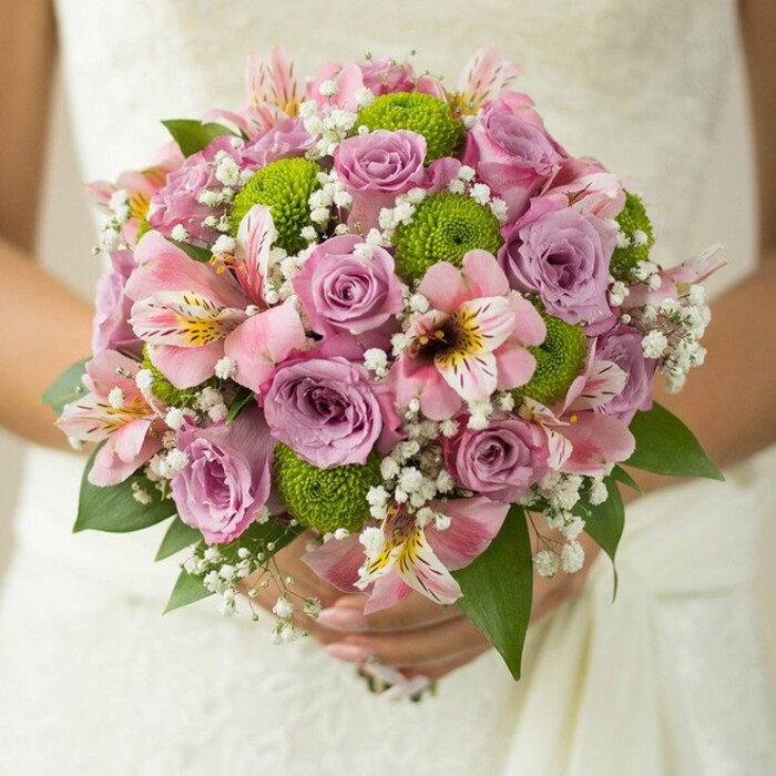 A breathtaking bridal bouquet rests gently in a bride's hands, the expertly arranged flowers standing out against the flowing white fabric of a wedding dress somewhere near Vauxhall's historic landmarks. The round, opulent arrangement is anchored by rose varieties in hues of pale pink and silvery lavender-some petals edged with morning dew-reflecting the romance of a riverbank sunrise. Popping through the pastel tones are crisp lime green button chrysanthemums, their lively spheres adding texture and modernity, much like the vibrant green spaces found near Oval or Robsart Street. Peruvian lilies with soft pink petals and unique speckled centers bring delicate fascination and a subtle fragrance, softening the arrangement with touches of grace. Interwoven clouds of tiny gypsophila fill out the bouquet, their white clusters providing airiness akin to light drifting through Vauxhall's open squares. The bride's fingers, adorned with a simple silver ring, cradle the neatly wrapped stems, illustrating a moment of calm anticipation. The entire bouquet embodies tenderness and hope, seamlessly blending classic beauty with contemporary flair-perfect for metropolitan weddings and inspired by the unique energy of Vauxhall. This floral creation is not just a feast for the eyes but an emotional experience, echoing the fresh beginnings celebrated right in the heart of London.