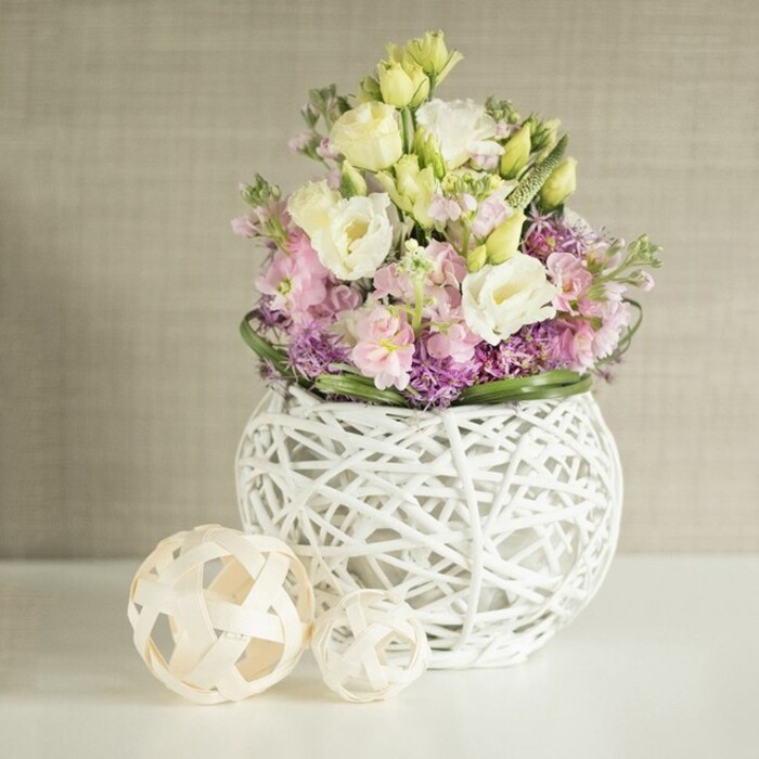 This inviting floral display, expertly crafted for the heart of Vauxhall, blends artistry with the soft romance of spring blooms. A generous bouquet of creamy white and pale yellow lisianthus-known for their elegant, ruffled petals-rise gently from a distinctive round vase, whose modern open-weave silhouette calls to mind the contemporary designs often seen in the galleries and artisan shops dotted across Vauxhall. Pink matthiola, or stock flowers, stretch upwards among the eustoma, infusing the bouquet with gentle height and an air of lush abundance. Sprays of vibrant purple filler-suggestive of aster or young allium-nestle at the base, their clusters creating a lively contrast against a circular band of deep, glossy green foliage. This visual medley finds unity in the vase, a sphere formed from intertwining rattan or painted twigs, lending both dimension and lightness that's artfully echoed by two decorative, woven spheres set to the left on the pristine white surface. The tones of beige and cream in the softly textured wall behind evoke a tranquil, sunlit room overlooking Spring Gardens or Vauxhall's riverside flats. The overall aesthetic is one of refined celebration, ideal for intimate wedding tablescapes or thoughtful springtime gifts for loved ones in Vauxhall. With each detail thoughtfully placed, the scene radiates calm elegance-a testament to the skill and passion of local florists, who curate same-day flower delivery across the Vauxhall neighbourhood.